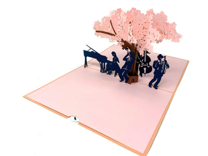 &quot;Musicians under blossom tree pop-up card: A three-dimensional card with an intricately crafted pop-up scene featuring charming musicians performing under a blossoming tree, creating a delightful and artistic setting.&quot;